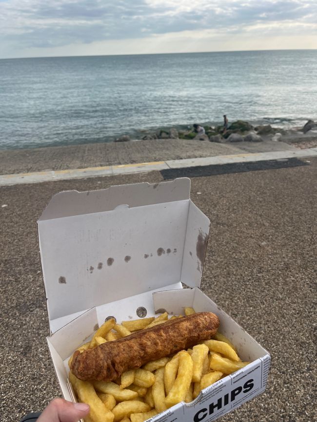A sausage in batter and chips all in a box being held in front of the sea